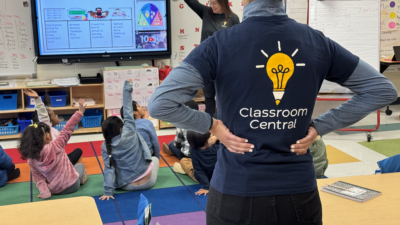 Students sitting on a carpet with their hands raised, engaged in learning, while a volunteer wearing a Classroom Central logo t-shirt assists
